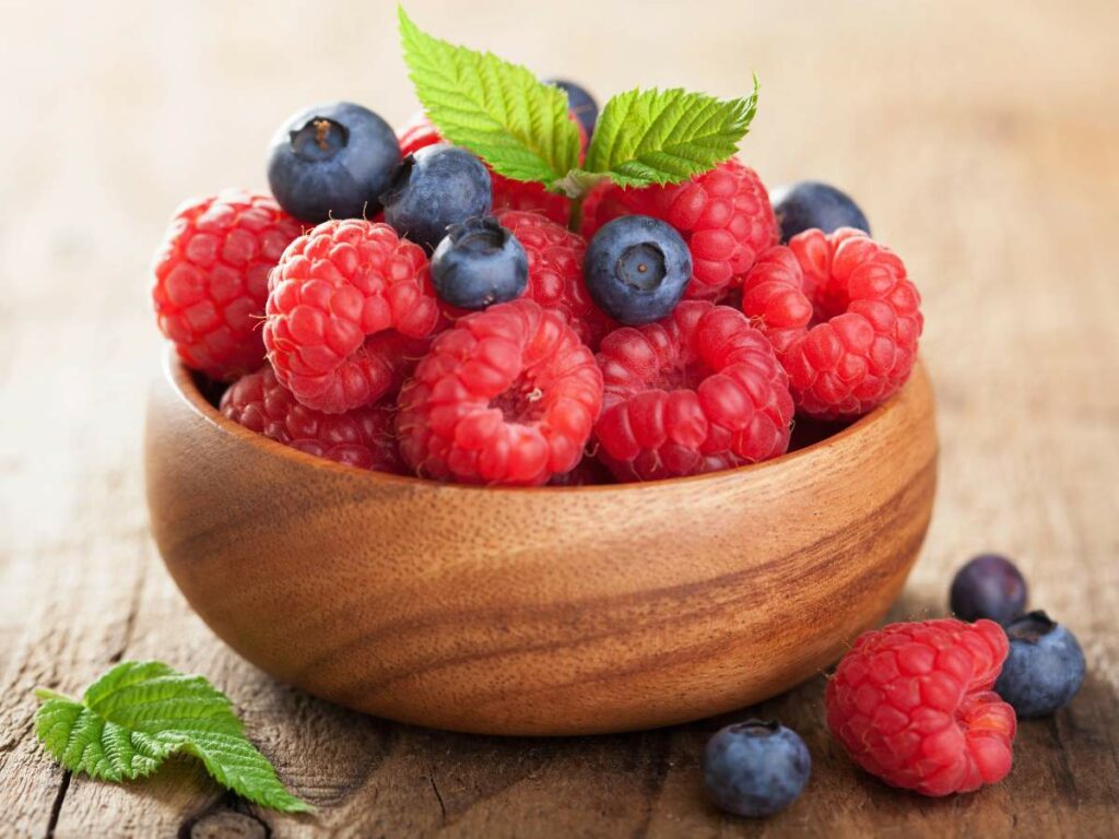 A wooden bowl filled with fresh raspberries and blueberries, with a few green leaves garnishing the top. The bowl is placed on a rustic wooden surface.