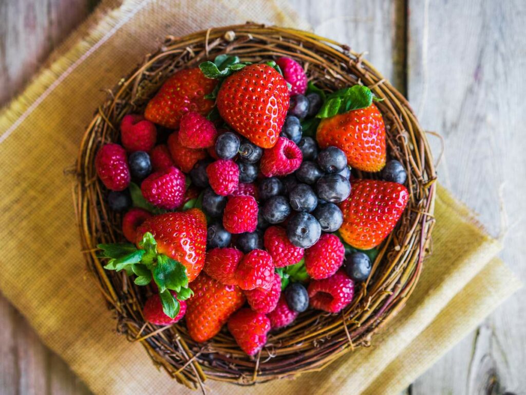 A rustic nest-like container filled with fresh strawberries, raspberries, and blueberries sits on a piece of burlap on a wooden surface.