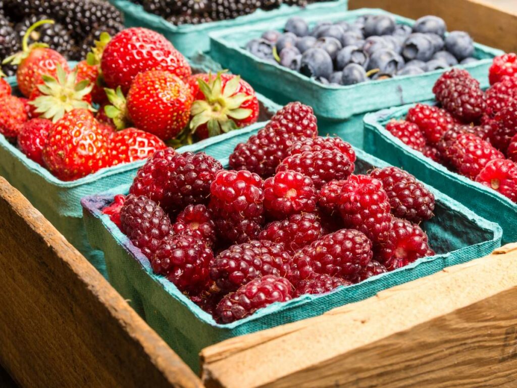 Various berries including strawberries, blueberries, blackberries, and raspberries are displayed in green cartons in a wooden crate at a market.