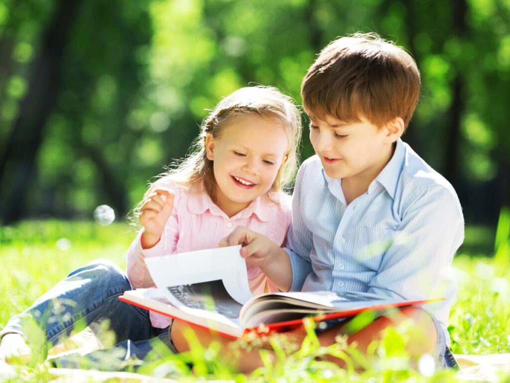 Two children sit on grass in a park, smiling and looking at a book together on a sunny day.