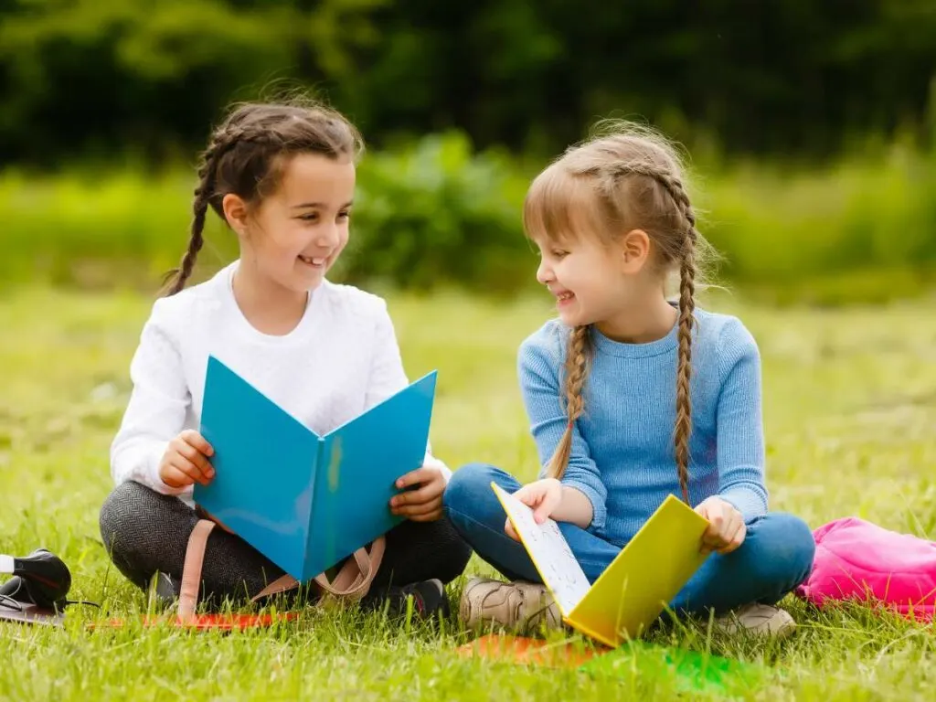 Two young girls with braided hair sit on grass, smiling and holding open books.