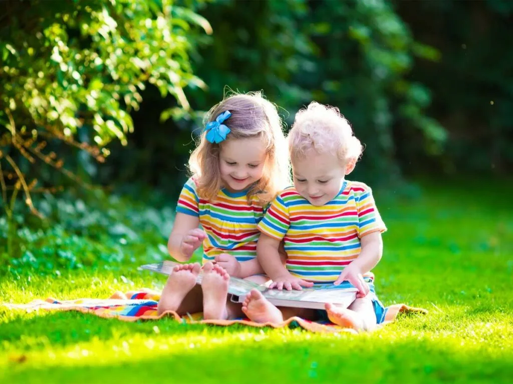 Two young children, wearing matching striped shirts, sit on a blanket on the grass, looking at a book together.