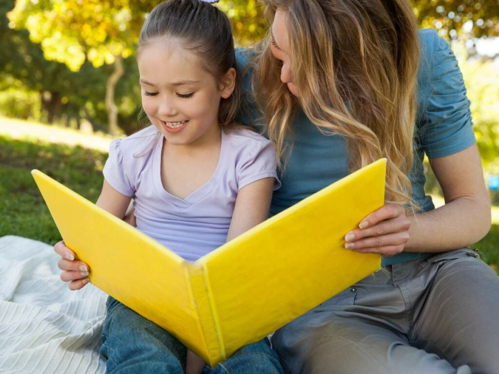 A woman and a young girl sit on a blanket outdoors, reading a large, yellow book together on a sunny day.