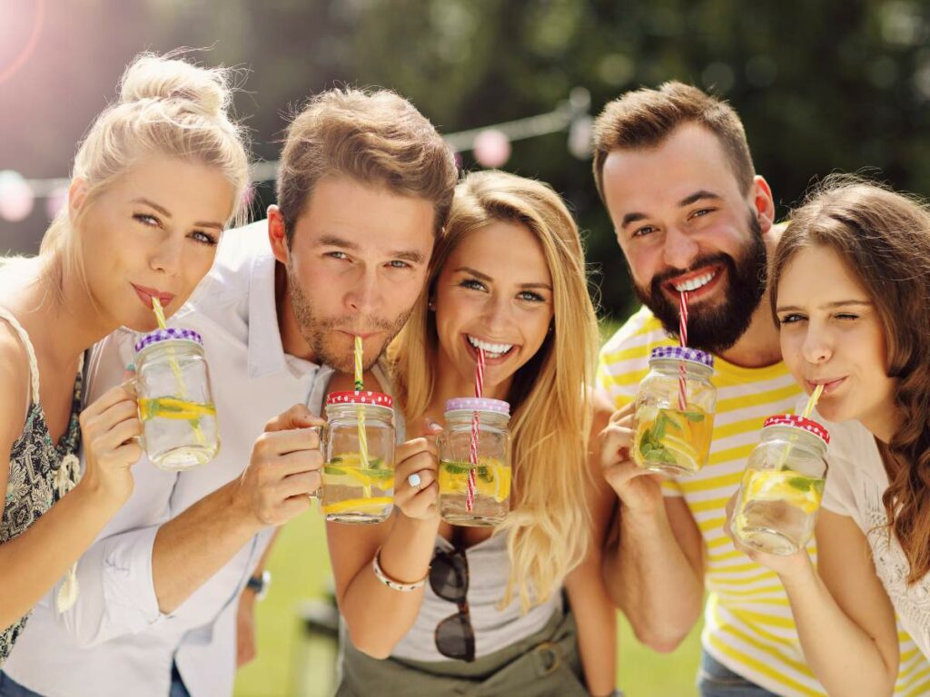 Five people smiling and drinking from mason jars with straws at an outdoor gathering.