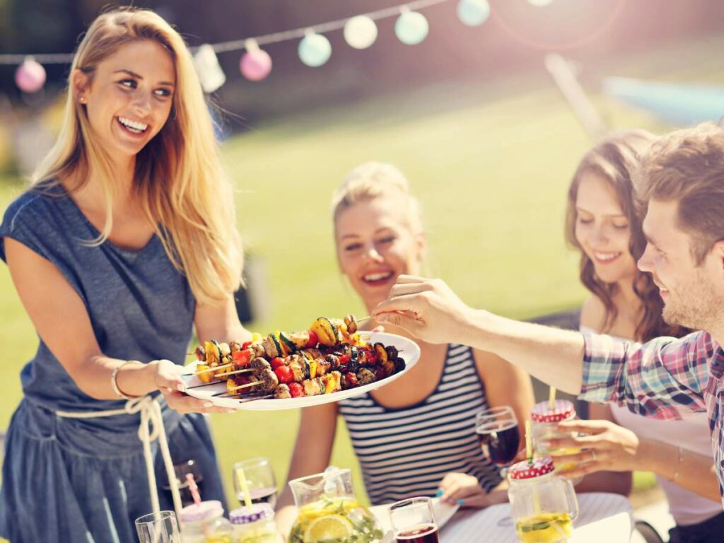 A woman serves skewered food to friends at an outdoor gathering. The group is sitting around a table with drinks and decorations in the background.