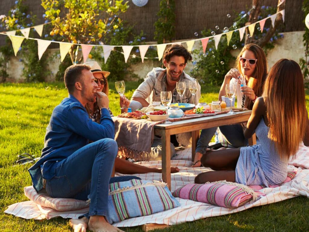 A group of people sitting on a picnic blanket in a garden, enjoying food and drinks around a low table, with colorful bunting in the background.