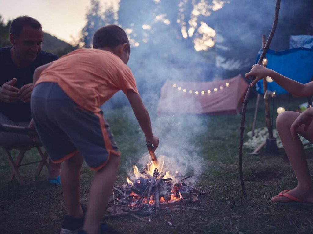 A child in a red shirt and grey shorts tends to a small campfire with adults nearby. Tents with string lights are in the background, set in a wooded area during twilight.