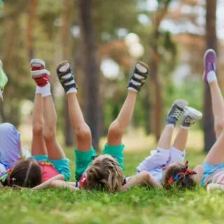 Five children lying on the grass with their legs raised in the air, wearing colorful clothes and shoes.