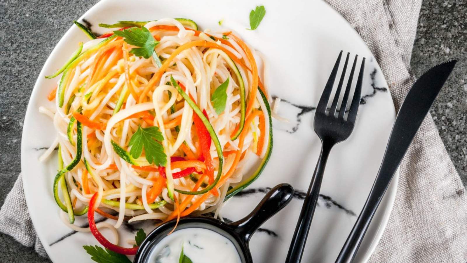 A plate of vegetable noodles garnished with parsley, served with a small bowl of sauce on a marble-patterned plate with a black fork and knife.