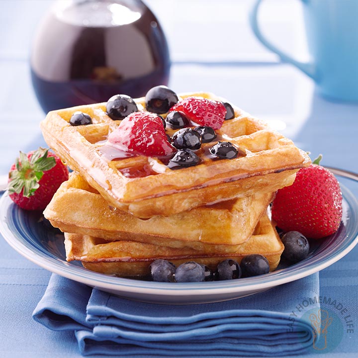 Plate of waffles topped with strawberries, blueberries, and syrup, with a dish of syrup in the background and a blue napkin underneath the plate.