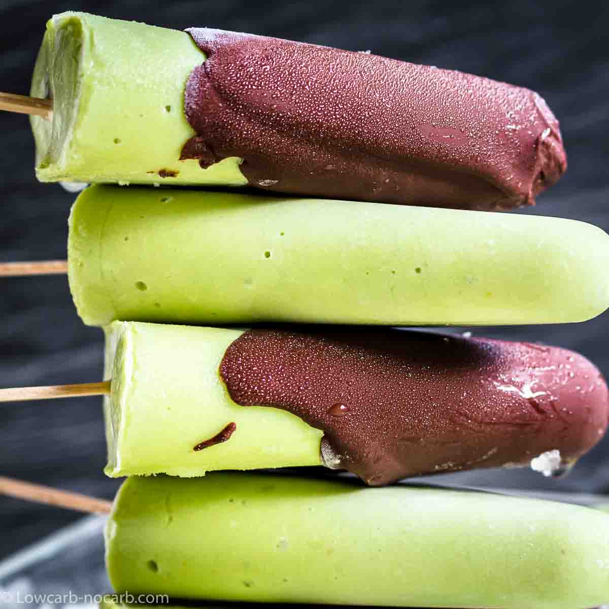 A stack of four green avocado popsicles with wooden sticks, two of which are partially coated in chocolate.