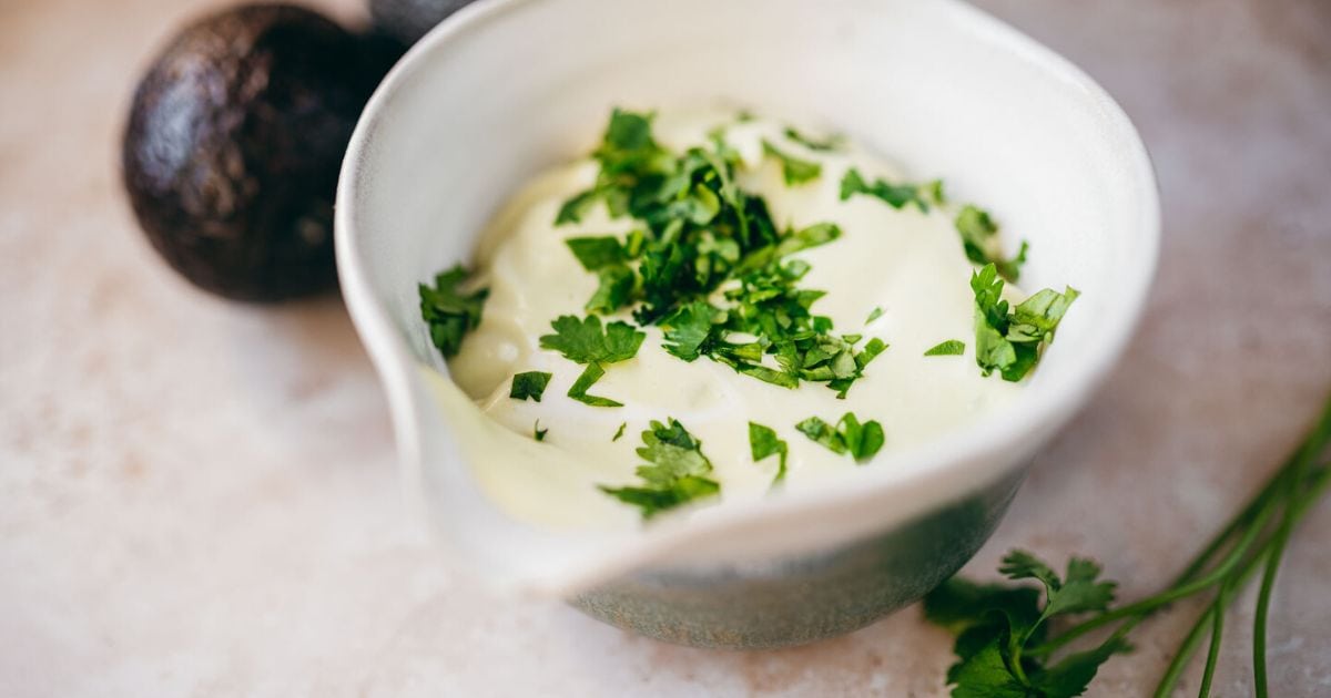 A bowl of creamy sauce garnished with chopped cilantro, with whole avocados and cilantro sprigs in the background.