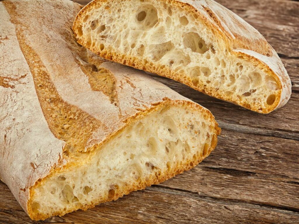 A sliced loaf of ciabatta bread with a crispy crust and airy interior is placed on a wooden surface.