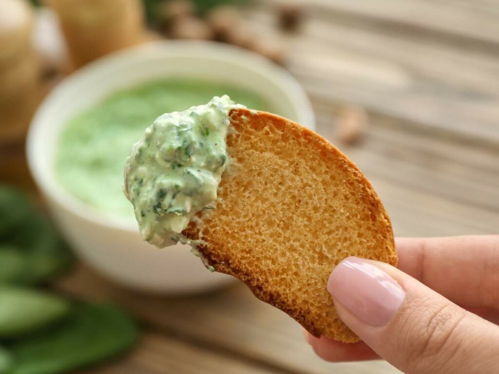 Close-up of a hand holding a slice of bread with a green dip on it.