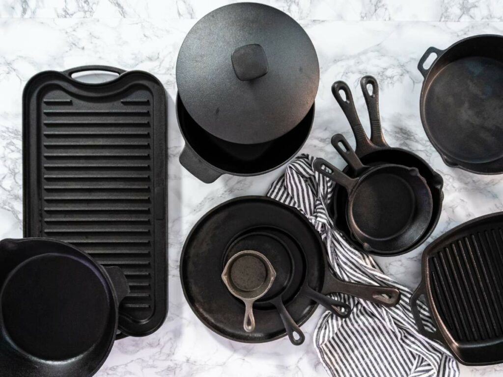 An assortment of black cast iron cookware, including skillets, a griddle, and a Dutch oven, arranged on a marble surface with a black and white striped cloth.