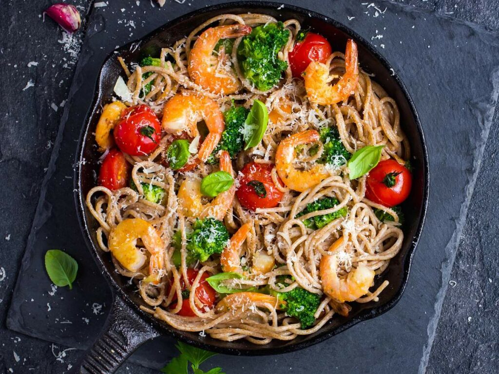 A skillet filled with shrimp pasta, broccoli, cherry tomatoes, and sprinkled parmesan cheese, garnished with fresh basil leaves.