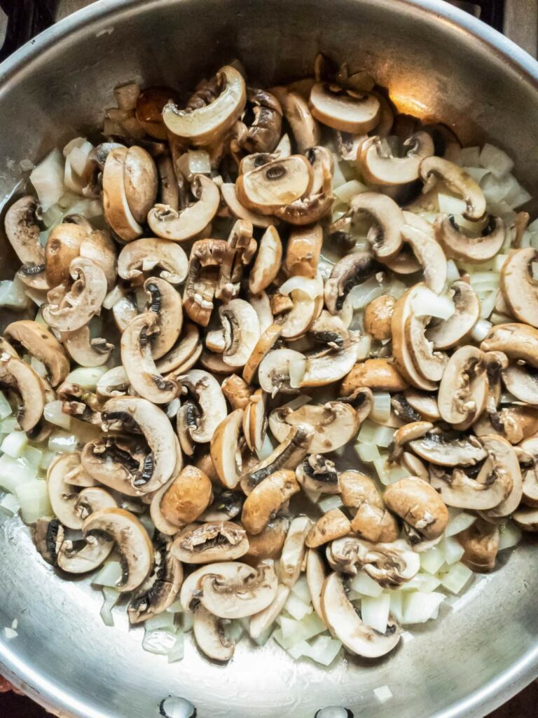 A metal pan filled with sliced mushrooms and diced onions being sautéed.