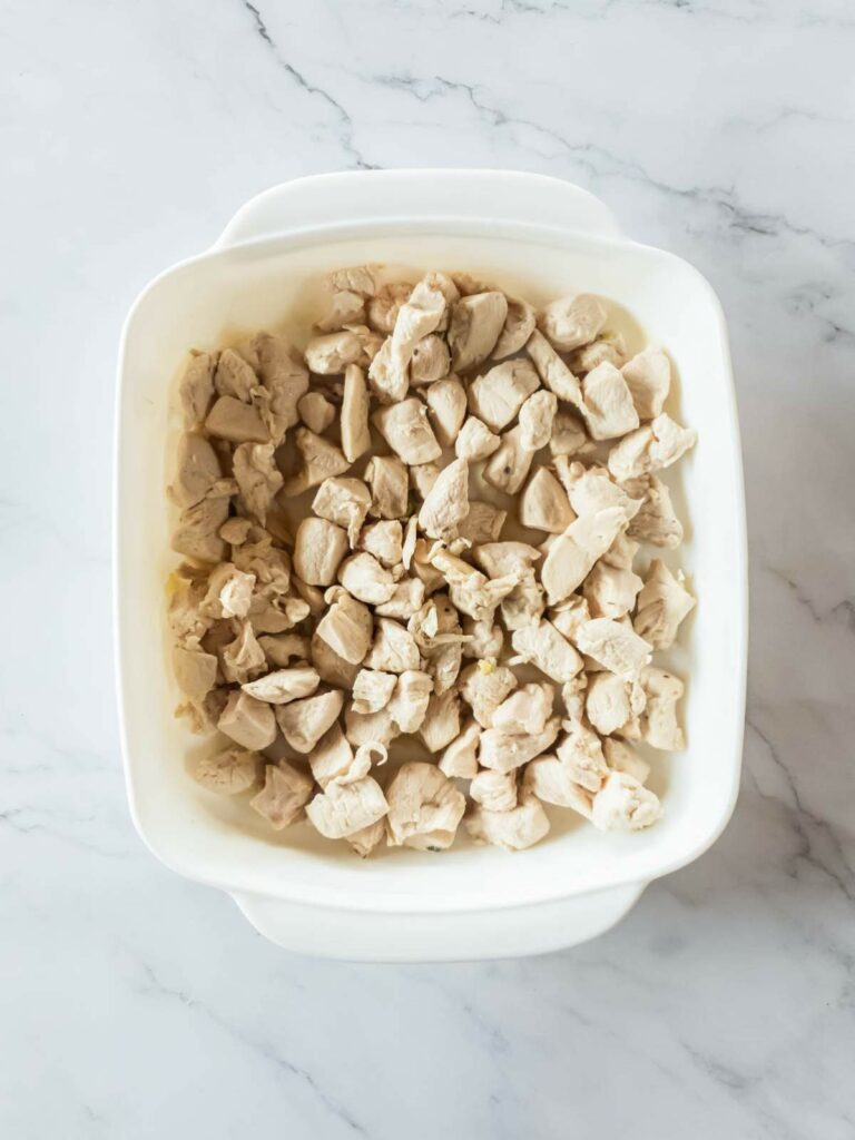 A white dish filled with chunks of cooked chicken on a marble counter surface.