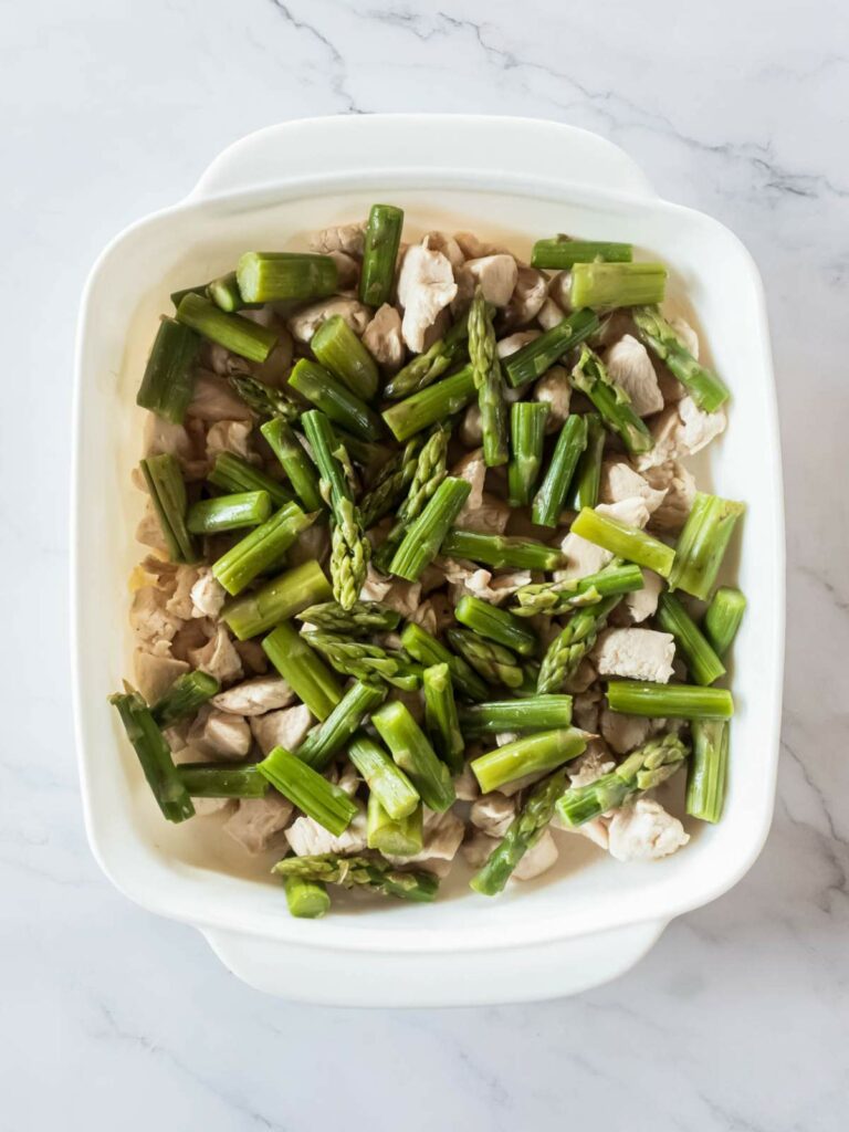 A white dish filled with pieces of cooked chicken and chopped asparagus on a marble surface.