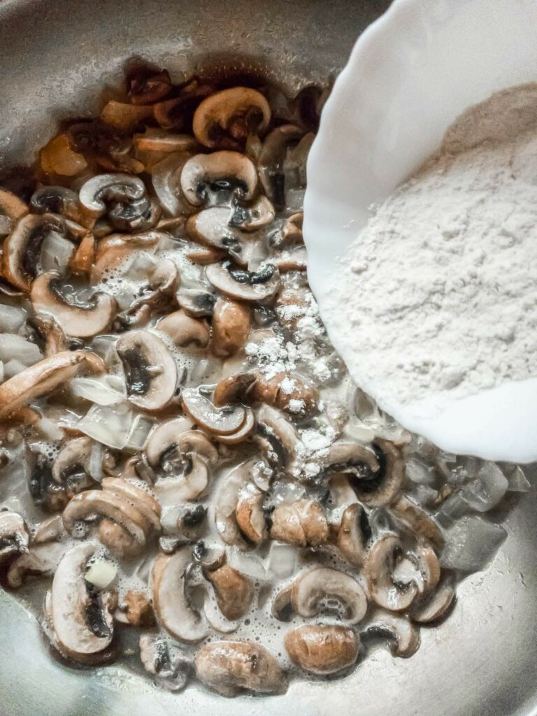 Sliced mushrooms and diced onions cooking in a pan with a bowl of flour being added.