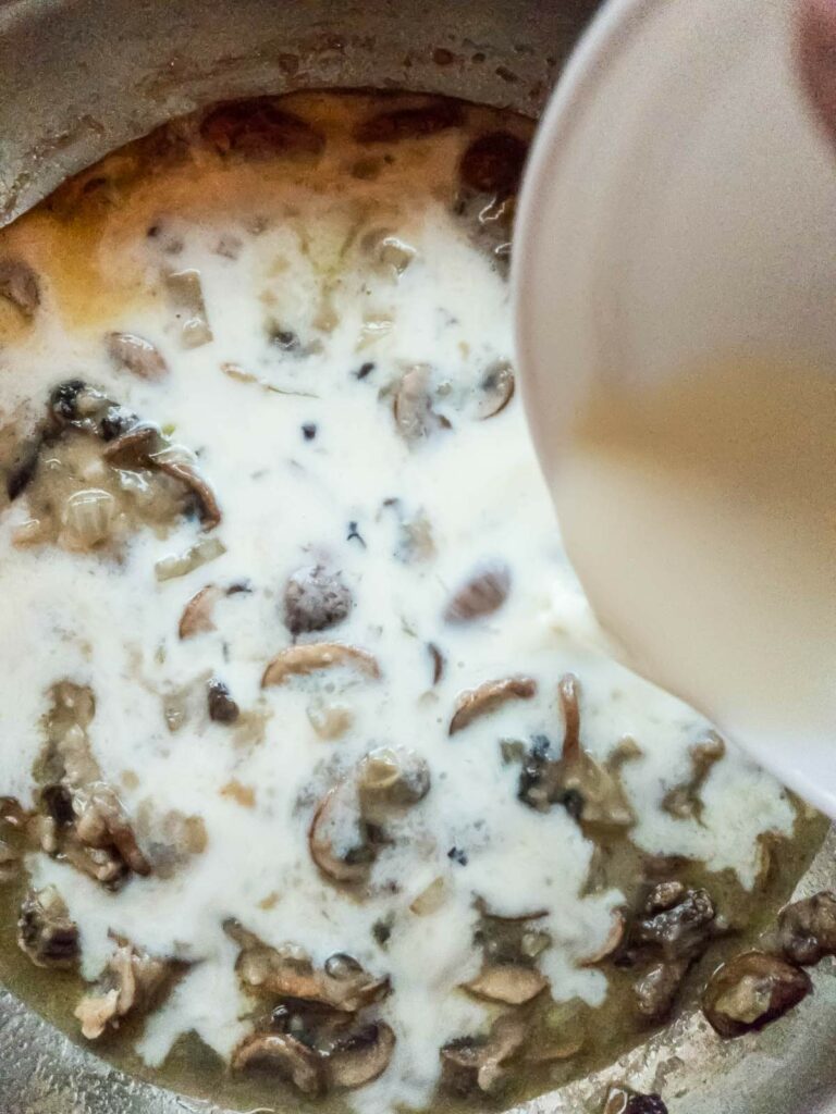 A person pours a creamy sauce into a pot containing mushrooms and other ingredients.