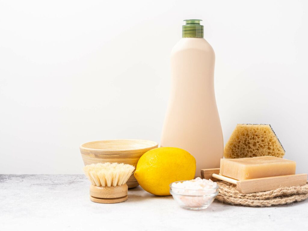 A set of personal care items on a white surface, including a bottle of lotion, a yellow lemon, soap, bath sponge, brush, bowl, and a small container of bath salts.