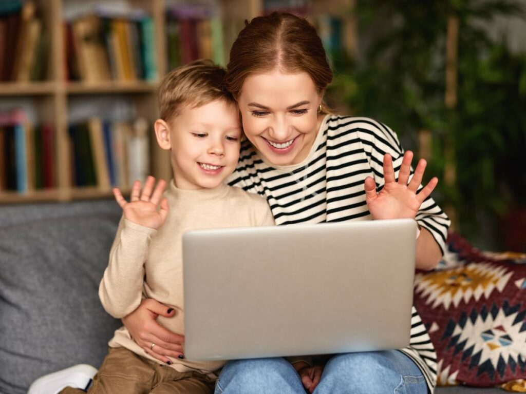 A woman and a young boy sitting together on a couch, smiling and waving at a laptop screen.