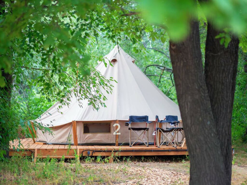 A beige triangular tent is pitched on a wooden platform surrounded by trees.