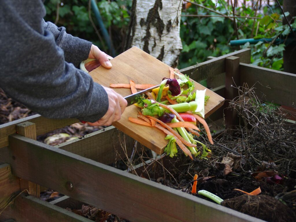 Person dumping vegetable scraps from a cutting board into a compost bin in a garden.