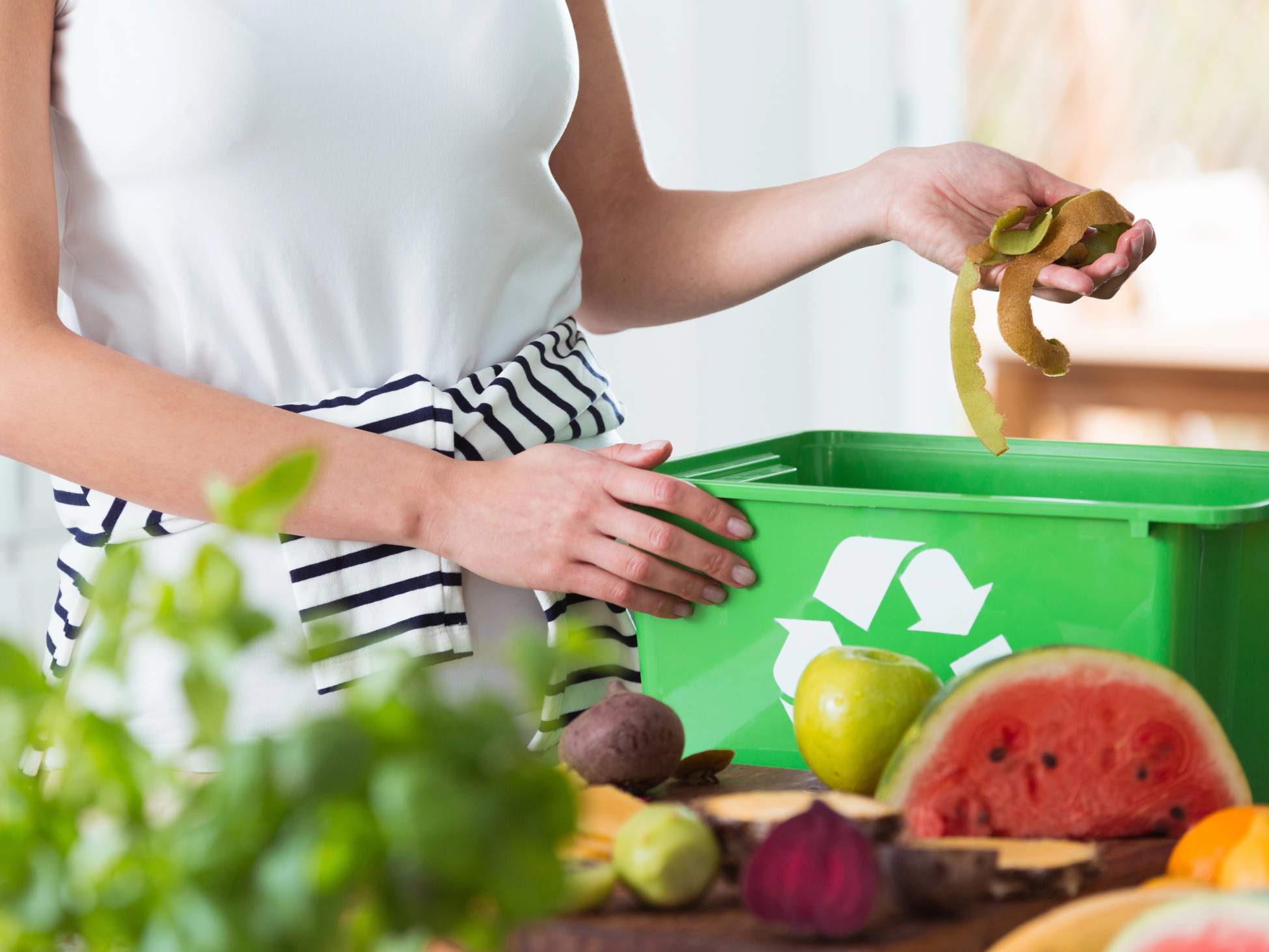 Person holding fruit peels over a green recycling bin in a kitchen with various fruits and vegetables on the counter.