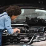 A mechanic works under the raised hood of a car, inspecting or repairing the engine.