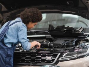 A mechanic works under the raised hood of a car, inspecting or repairing the engine.