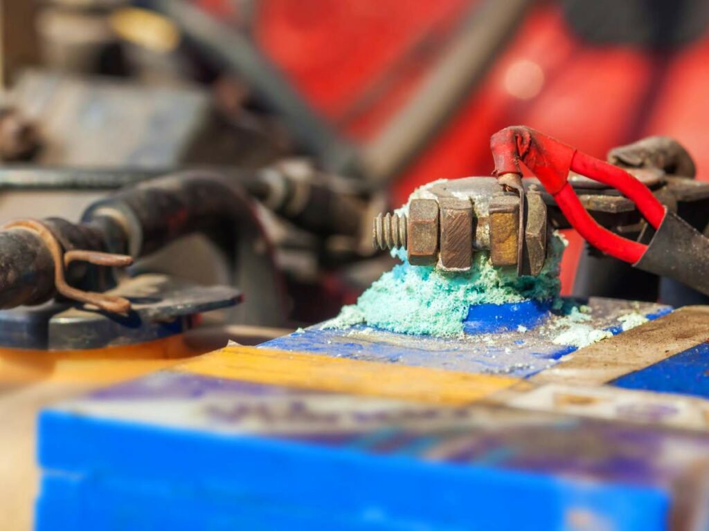 Close-up of a car battery terminal showing corrosion with blue-green deposits around the metal connectors and cables.