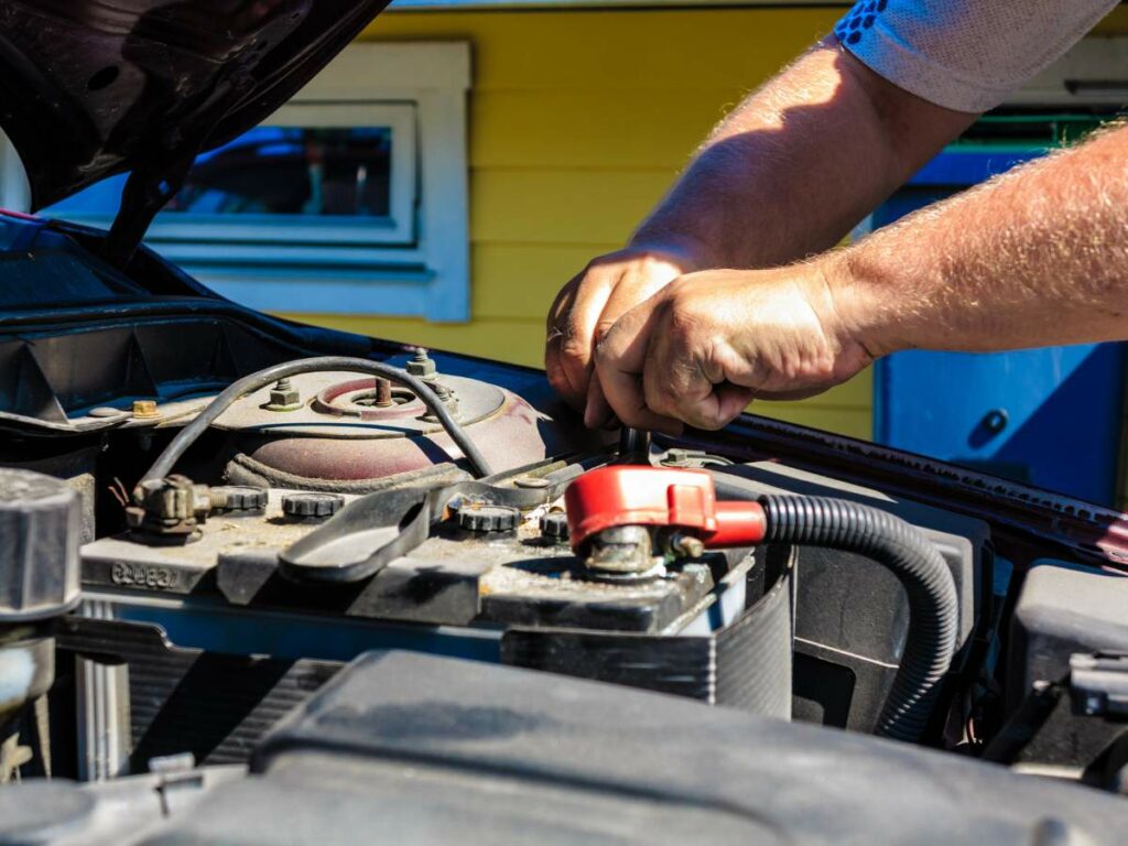 A person works on the engine of a vehicle with the car's hood open, using tools to adjust or repair components near the battery.