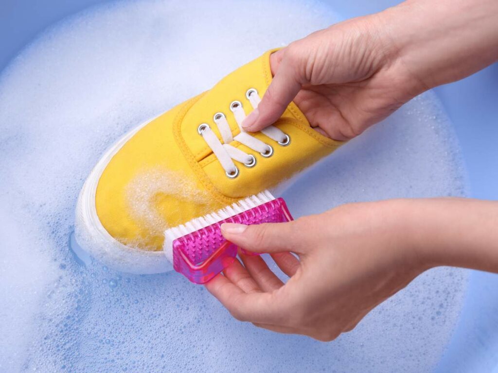 Hands scrubbing a yellow sneaker with a pink brush in soapy water.