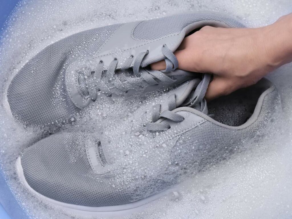 A hand is washing a pair of grey sneakers in soapy water.