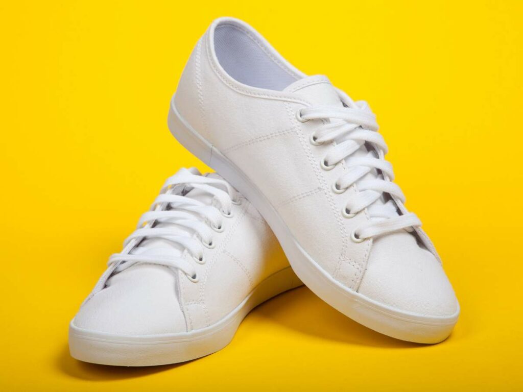 A pair of white canvas sneakers with laces, one leaning on top of the other, placed against a bright yellow background.