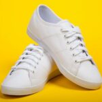 A pair of white canvas sneakers with laces, one leaning on top of the other, placed against a bright yellow background.