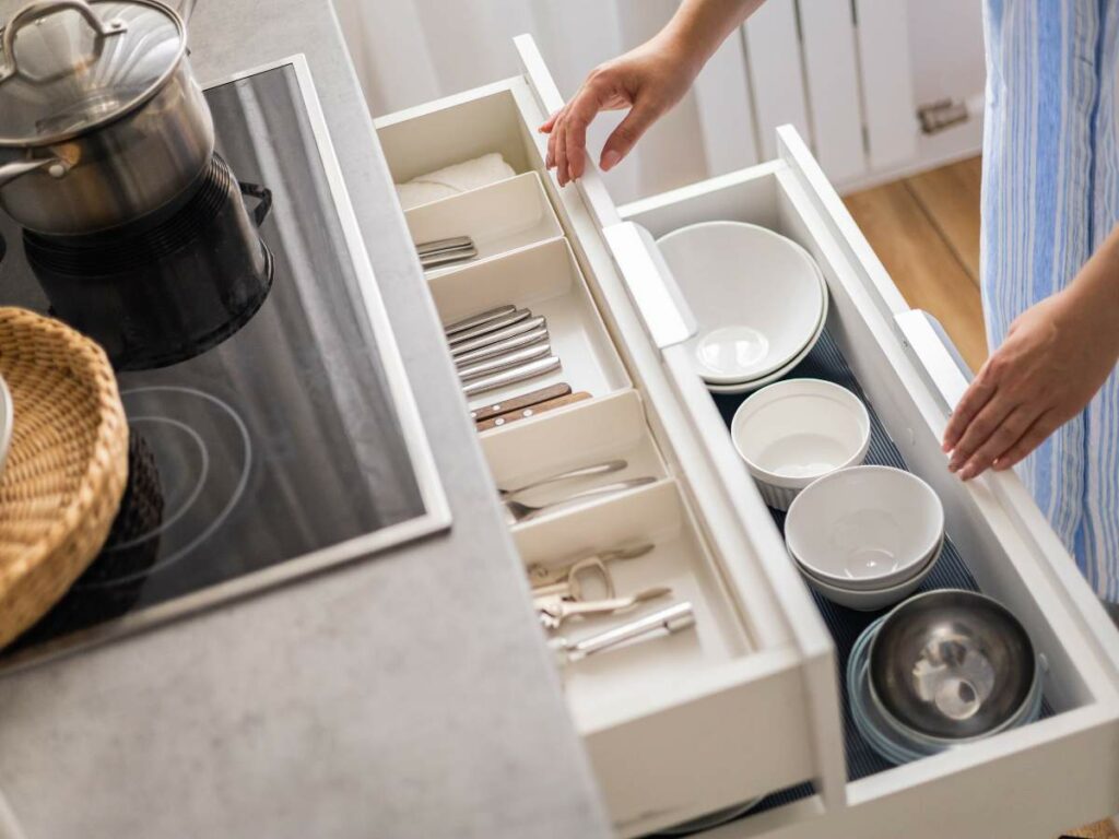 Person opening kitchen drawers containing cutlery and bowls near a stovetop with a pot and a wicker basket.