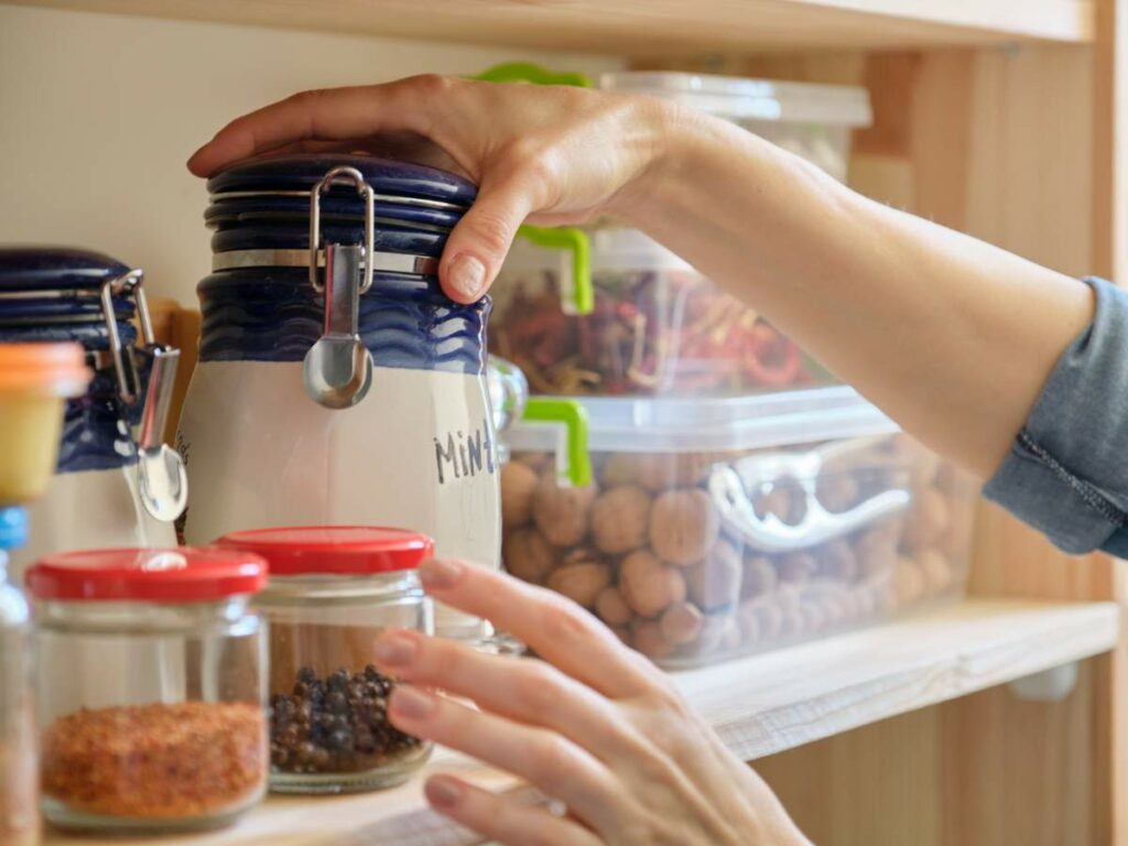 Hand reaching for a labeled jar on a pantry shelf, surrounded by various jars and containers.