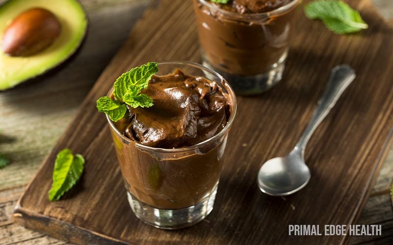 Two glasses of chocolate avocado mousse garnished with mint leaves sit on a wooden board.