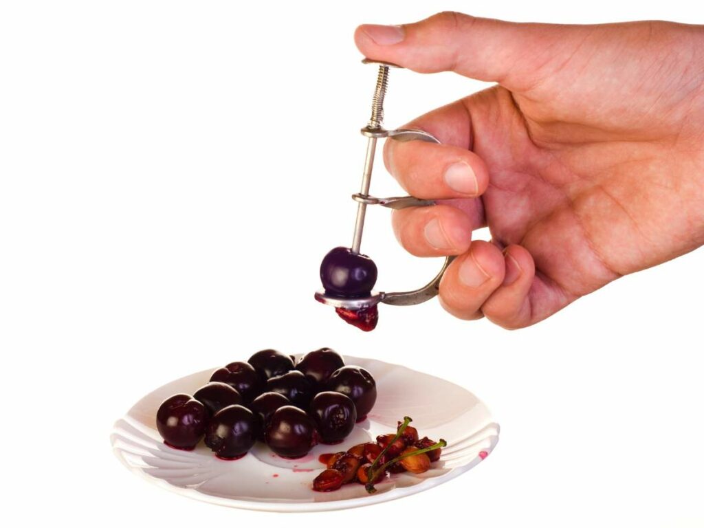 A hand uses a cherry pitter to remove pits from dark cherries on a white plate.