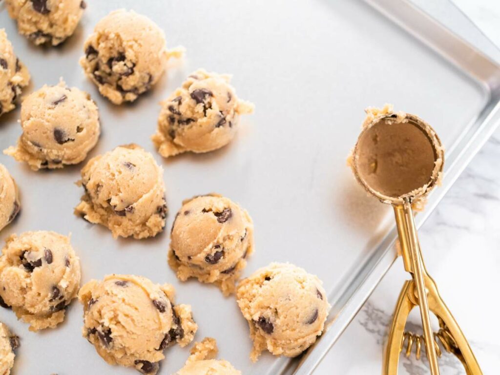 A baking tray with several balls of chocolate chip cookie dough and a cookie scoop.