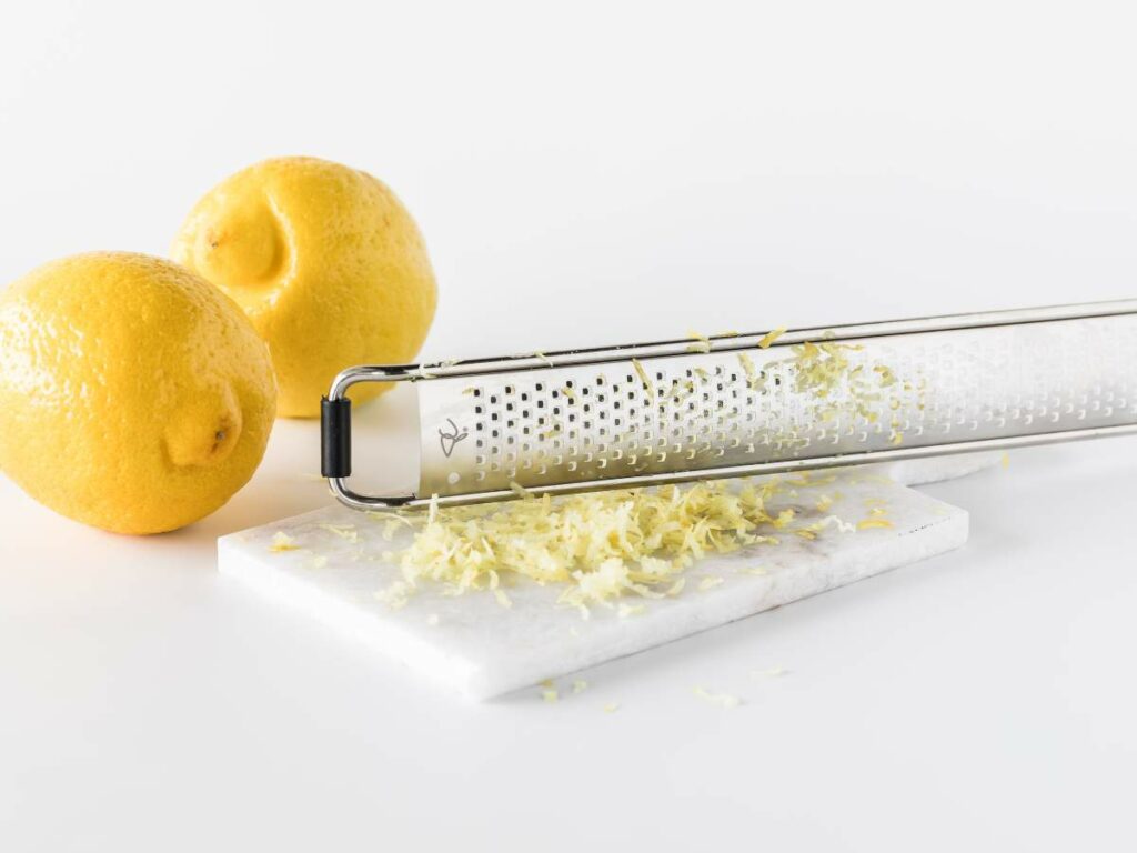 Two whole lemons and a zester with lemon zest on a marble slab.