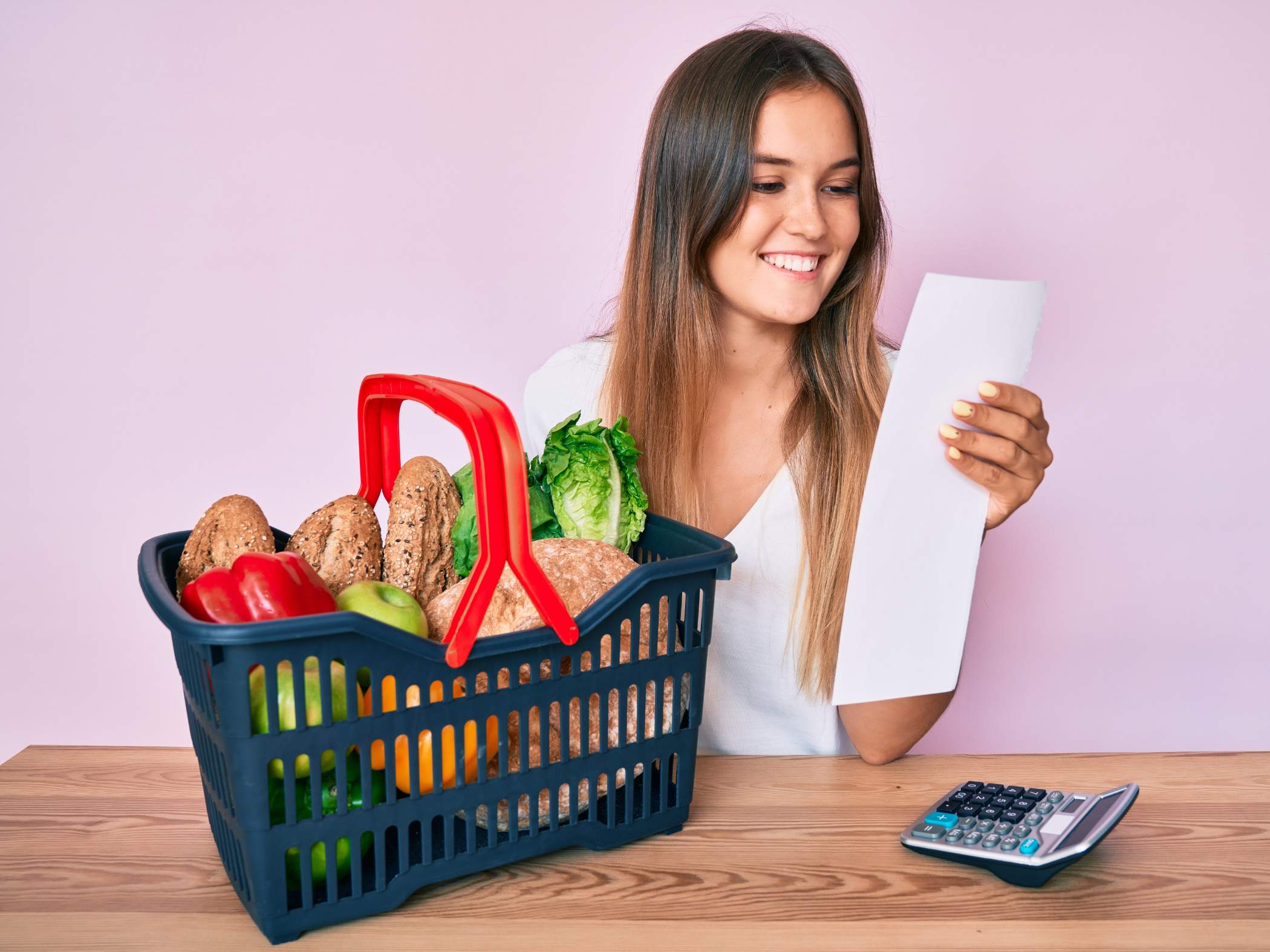 A smiling woman looks at a receipt while sitting at a table with a shopping basket full of groceries and a calculator.