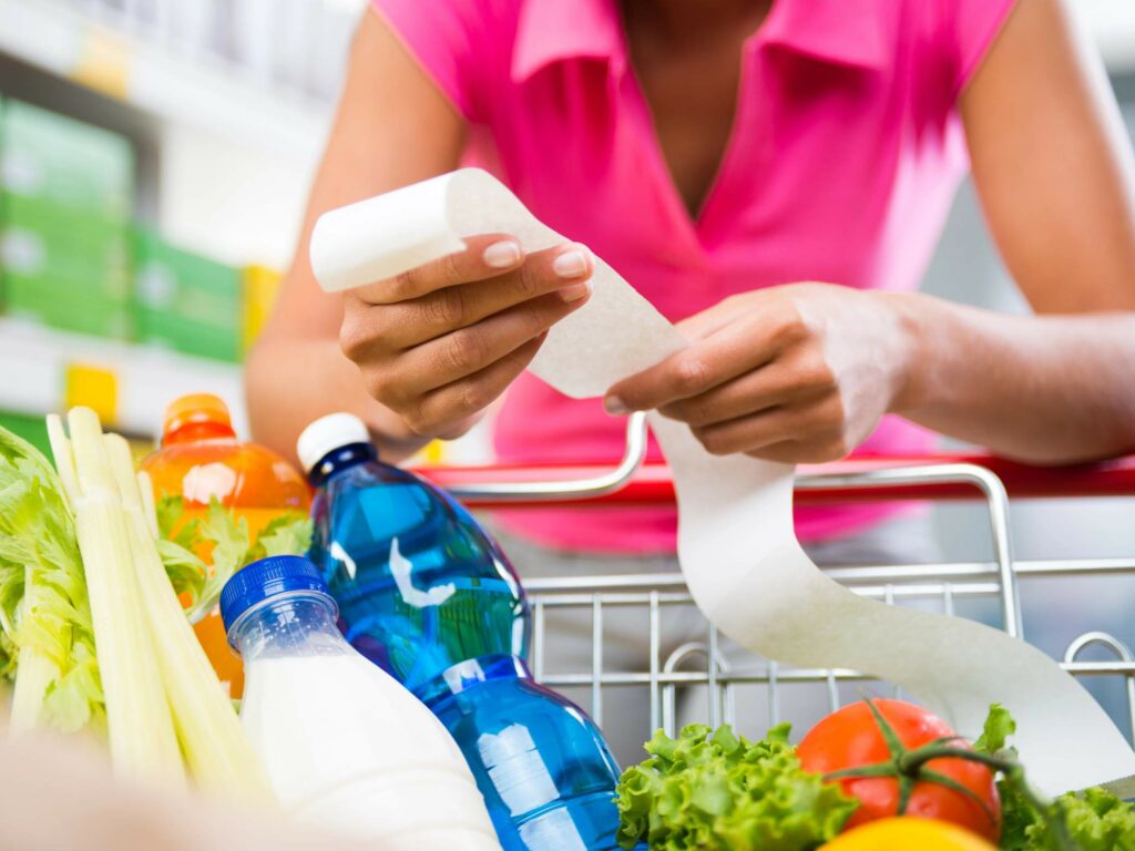 A person reviews a long receipt while shopping. The grocery cart is filled with a variety of items.