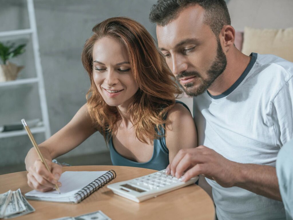 A woman with red hair writes in a notebook while a man with a beard uses a calculator at a table.