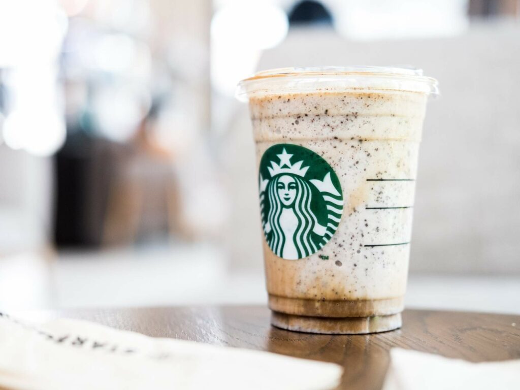 A plastic cup filled with a blended coffee beverage topped with whipped cream, bearing the Starbucks logo, sits on a wooden table.