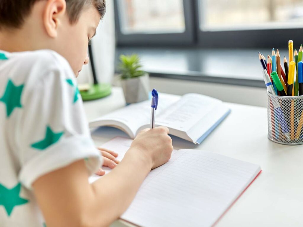 A young boy is sitting at a desk writing in a notebook with an open book in front of him.