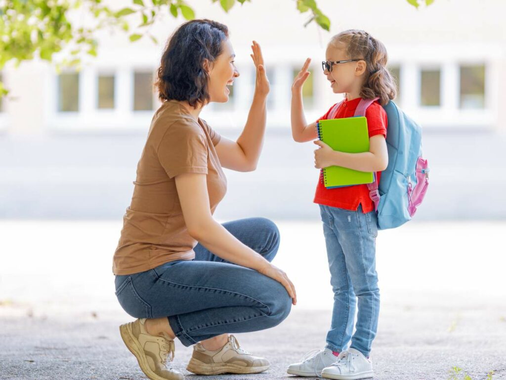 An adult and a child wearing a backpack and holding a notebook exchange a high-five outdoors.